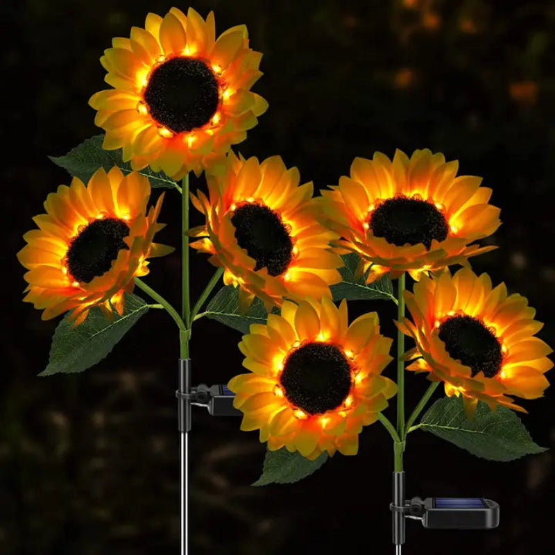 Six artificial sunflowers with dark centers and warm yellow glowing petals are shown against a dark background, likely a garden at night. Each flower is mounted on a stem with green leaves, and some have small black solar panels attached near the base, indicating they are solar-powered lights. The illumination creates a bright and cheerful display.