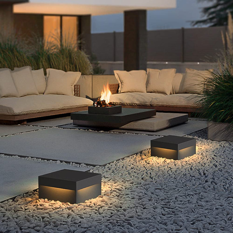 Three modern, square outdoor lights with dark tops and glowing warm-toned panels, illuminating a patio with white pebbles, lounge seating, and a fire pit at dusk.