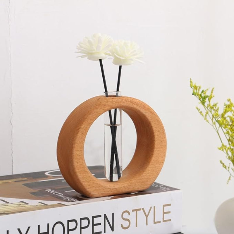 A light wooden circular vase holds a clear glass test tube with two artificial white flowers. The vase sits on a book titled "Hoppen Style".