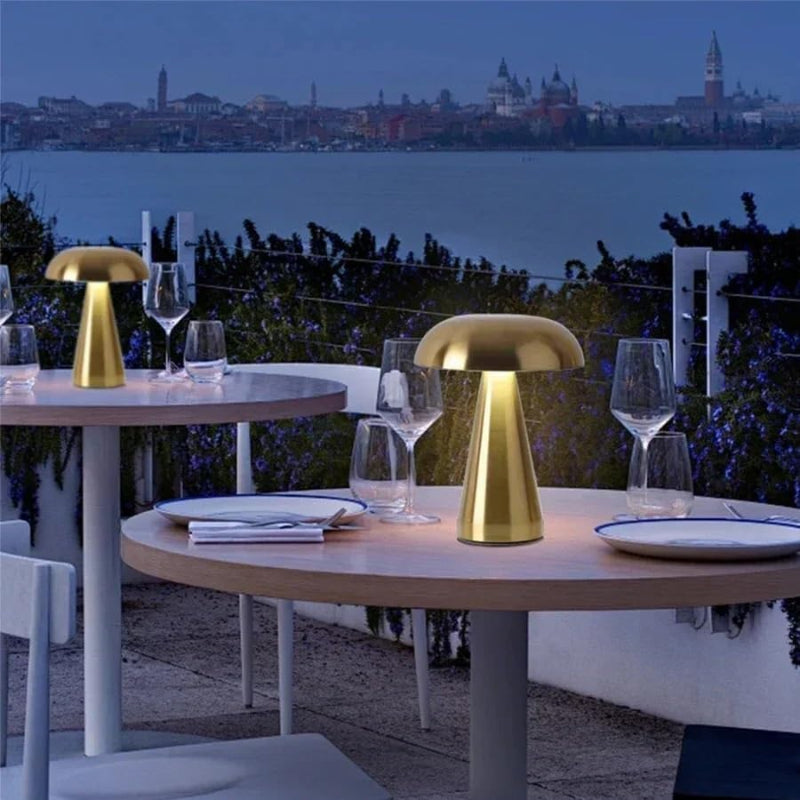 Two round tables are set for dining on an outdoor patio at dusk, each illuminated by a gold, mushroom-shaped, cordless lamp casting a warm glow on the white plates and wine glasses. The patio overlooks a body of water with a distant cityscape visible, creating a serene and inviting atmosphere for an evening meal.