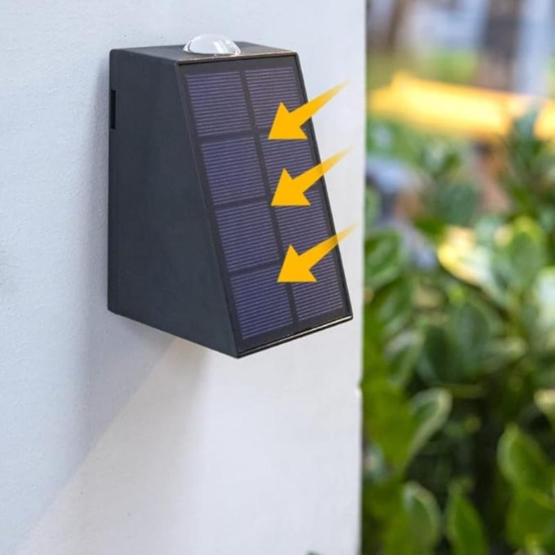 A small, dark, rectangular solar wall light is mounted on a light-colored surface. The light has a tilted solar panel with visible grid lines on its front. Three yellow arrows point towards the solar panel. Green foliage is visible in the blurred background.