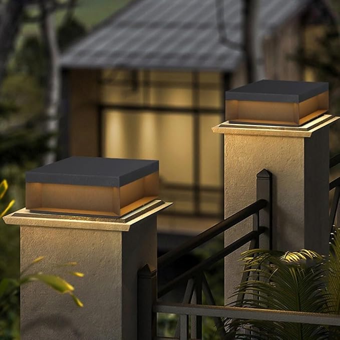 Two modern, square outdoor lights with dark tops and glowing warm-toned panels, mounted on top of light-colored gate pillars at dusk.