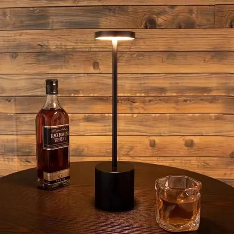 A sleek, cordless LED table lamp with a black finish, featuring a cylindrical base, a slender stem, and a circular light panel emitting a warm glow, situated on a dark wooden table next to a partially filled glass of amber liquid and a bottle of whiskey, against a rustic wooden plank wall.
