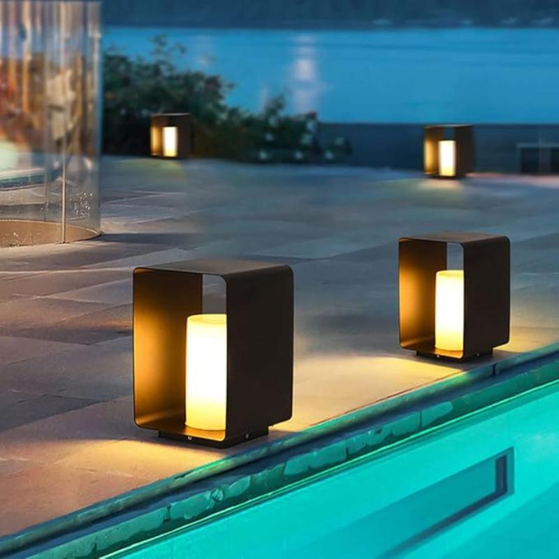 Four modern, dark-framed outdoor lanterns with glowing cylindrical lights inside, illuminating a patio area near a pool at dusk.