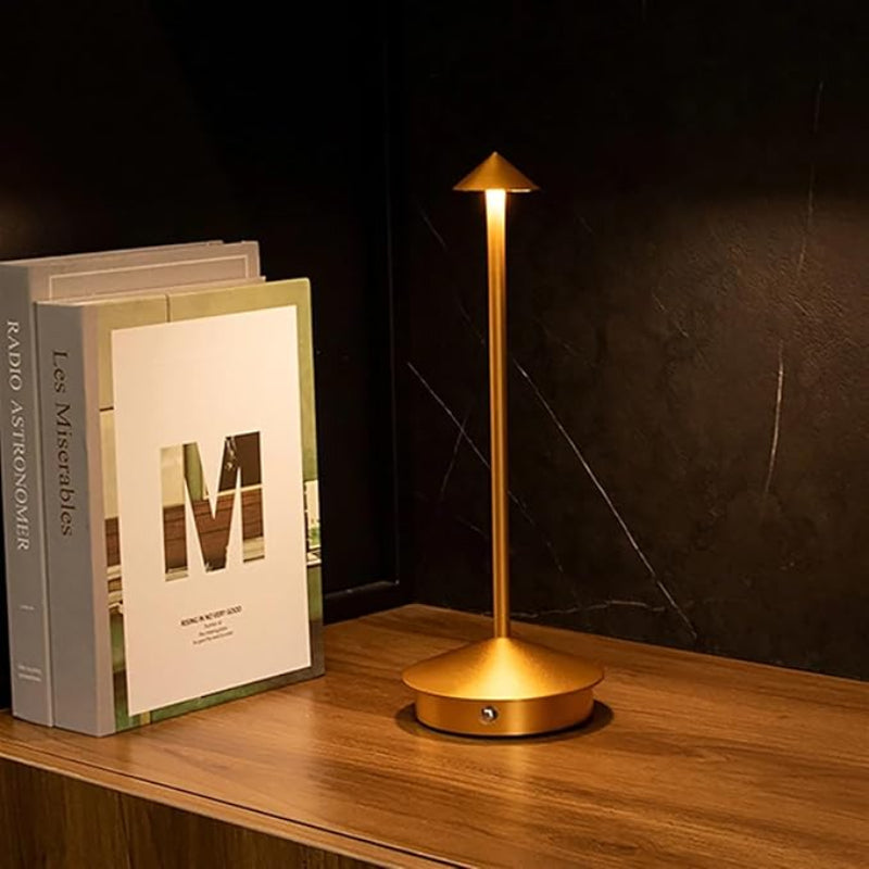 A lit, slender gold cordless lamp with a pointed shade stands on a wooden shelf next to two books. The background is dark.