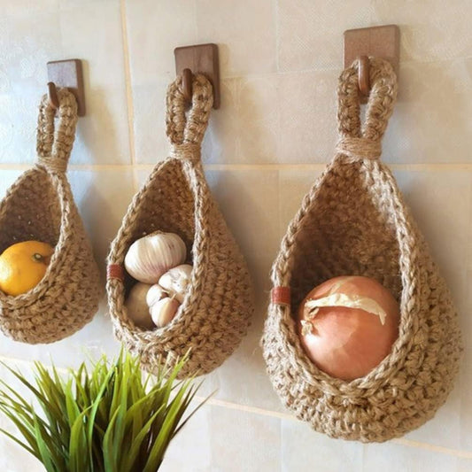 Three small, woven hanging baskets on a tiled wall, each holding a lemon, garlic, and an onion respectively. A potted plant is visible at the bottom.