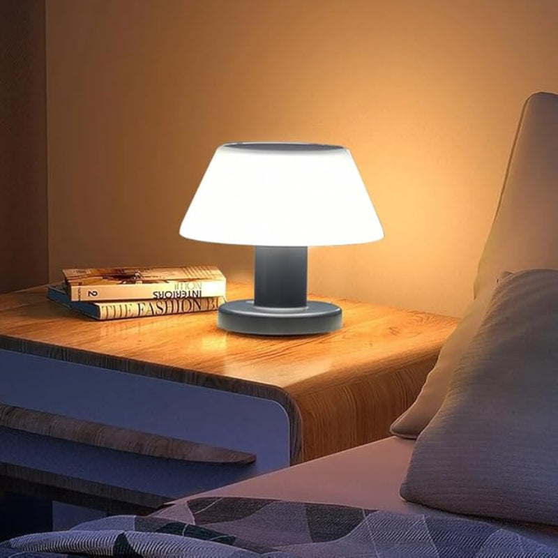 A small, modern table lamp with a white, slightly tapered lampshade and a dark, cylindrical base emits a soft glow on a wooden nightstand. Two books are stacked next to the lamp. Part of a bed with light-colored pillows and bedding is visible to the right. The warm light from the lamp illuminates the scene in a dimly lit room.