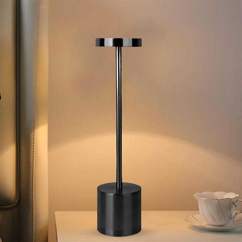 A sleek, cordless LED table lamp with a black finish, featuring a cylindrical base, a slender stem, and a circular light panel with a subtle glow, resting on a light-colored nightstand next to a delicate white teacup and saucer. The background shows a softly lit wall and part of a headboard.