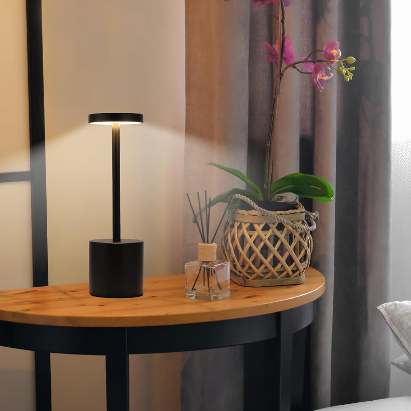 A cordless LED table lamp with a black cylindrical base, a slender stem, and a circular light panel emitting a soft glow, placed on a round wooden side table with black legs. The table also holds a square glass diffuser with reeds and a woven pot containing an orchid plant. The background features a window with sheer curtains and a glimpse of a bed.
