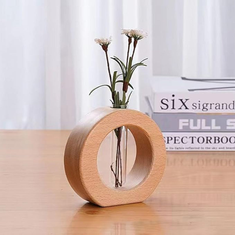 A light wooden circular vase holds a clear glass test tube with three small white flowers and green leaves. The vase sits on a light wooden surface in front of a stack of books.