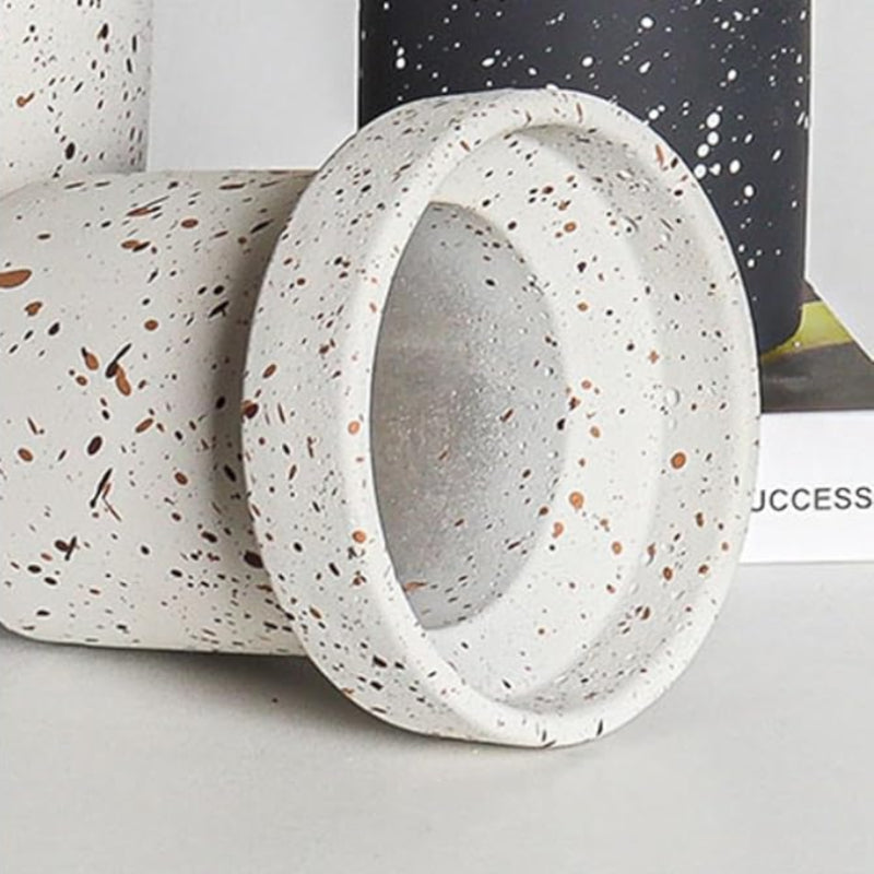 A close-up shows the rim and interior of a small, cylindrical white cement plant pot with brown and black speckles. The pot is lying on its side on a white surface. Part of another pot with a black and white speckled finish is visible in the upper right corner.