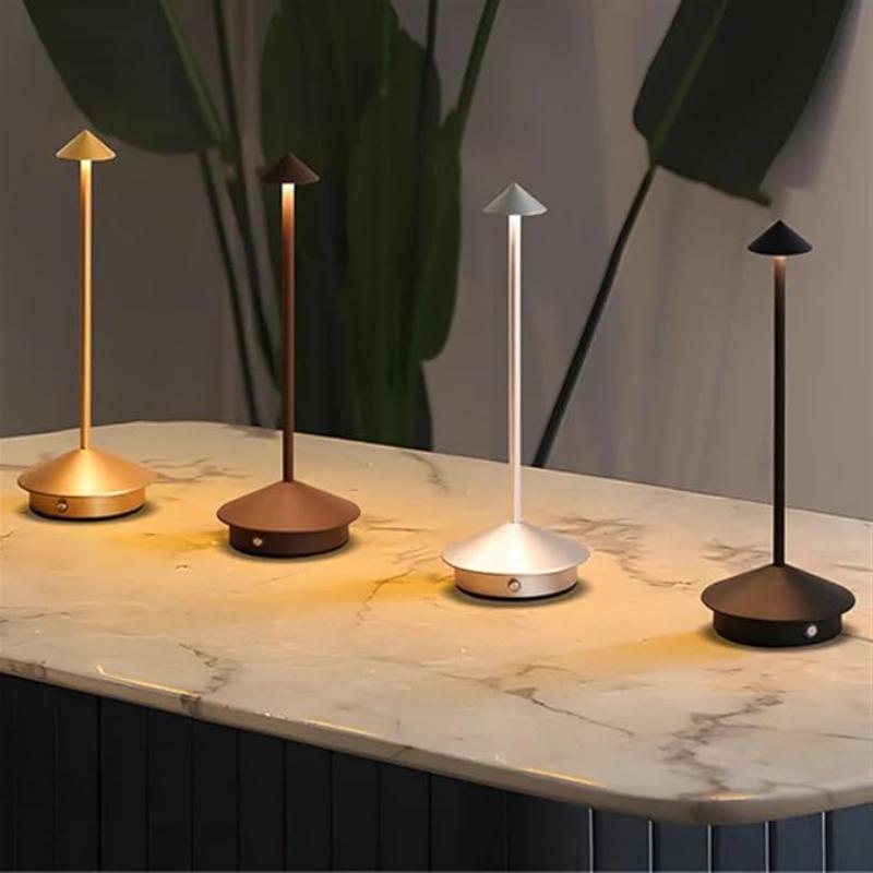 Four cordless table lamps in gold, bronze, silver, and black, each with a slim stem and a pointed shade, are illuminated on a marble-like countertop. Large green leaves are visible in the blurred background.