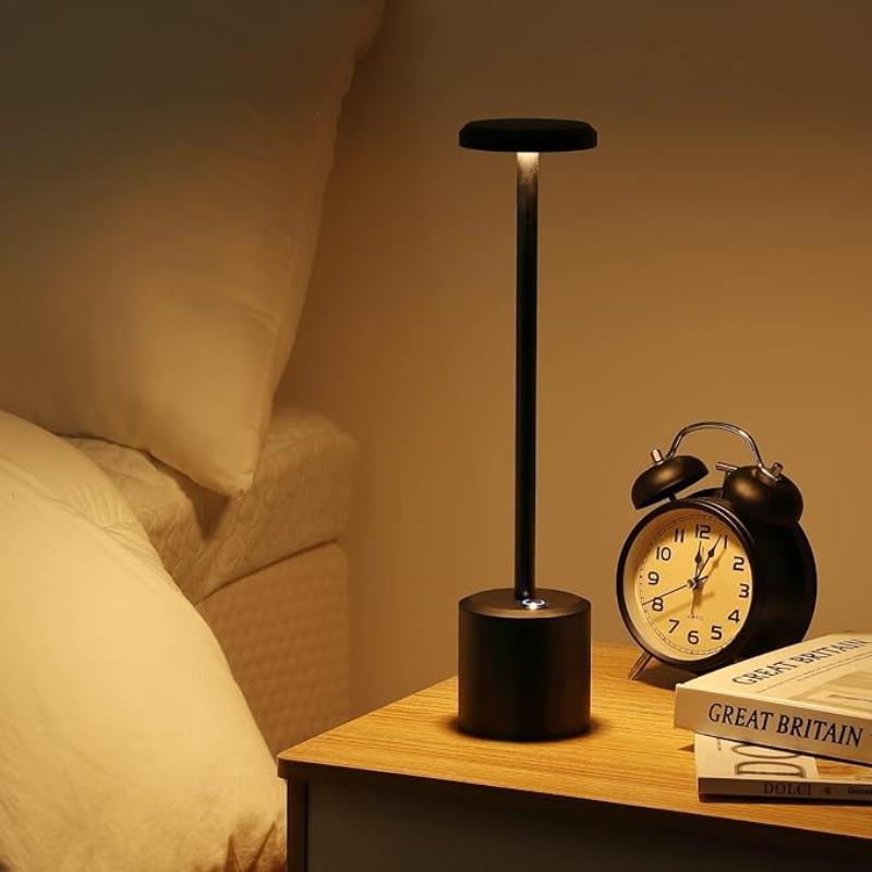 A cordless LED table lamp with a black cylindrical base, a slender stem, and a circular light panel emitting a soft glow, sitting on a wooden nightstand next to a black analog alarm clock and a stack of books near a bed with white linens.