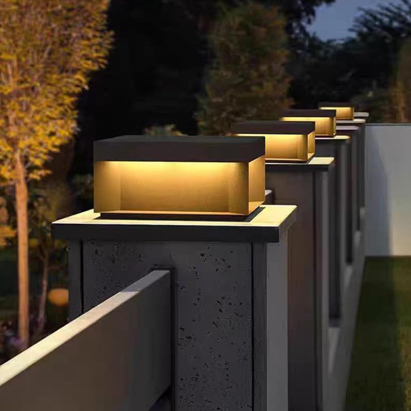 A series of contemporary, rectangular outdoor lights emitting a warm light, mounted on top of gray fence posts, lining a pathway at night.
