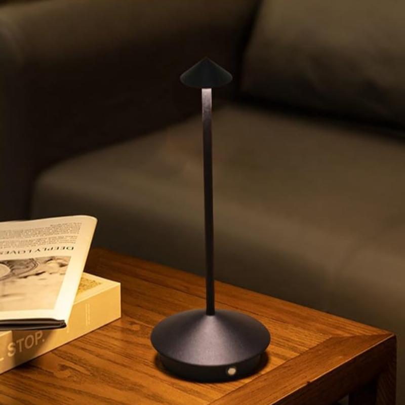 A small, cordless black table lamp with a pointed shade illuminates a wooden nightstand. Two books are stacked next to it. A blurred sofa is in the background.