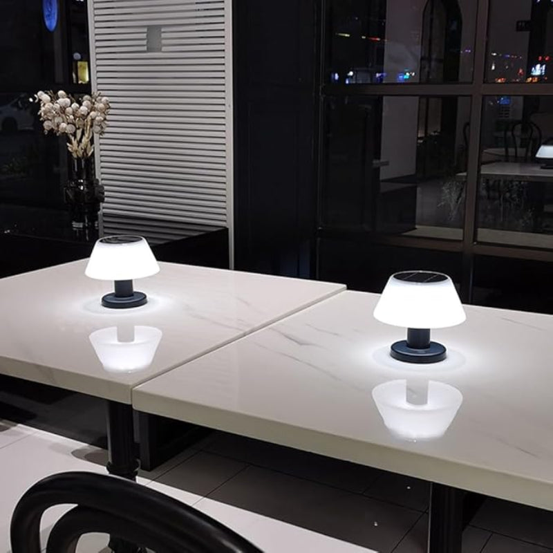 Two small, solar-powered table lamps with white, slightly tapered shades and dark bases illuminate two adjacent white tables in a dimly lit indoor setting, possibly a restaurant. Reflections of the lamps are visible on the table surfaces. A dark vase with dried flowers is on the left table. Large windows show blurred city lights at night in the background.