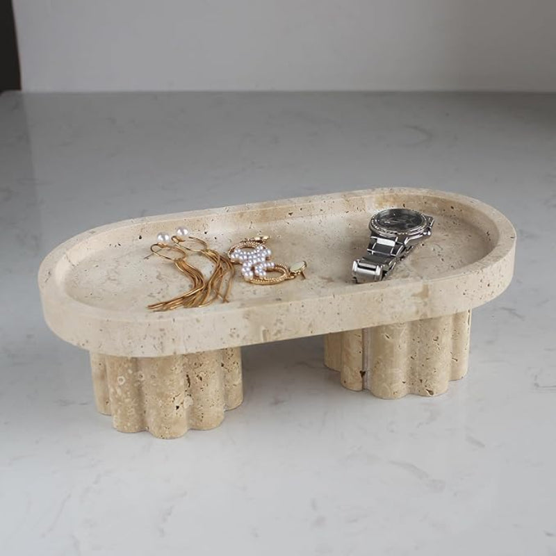 An oval, textured light-colored travertine tray with two fluted pedestal legs rests on a light marble countertop. The tray holds a silver wristwatch, a gold-toned necklace with pearl accents, and a pair of gold-toned earrings.