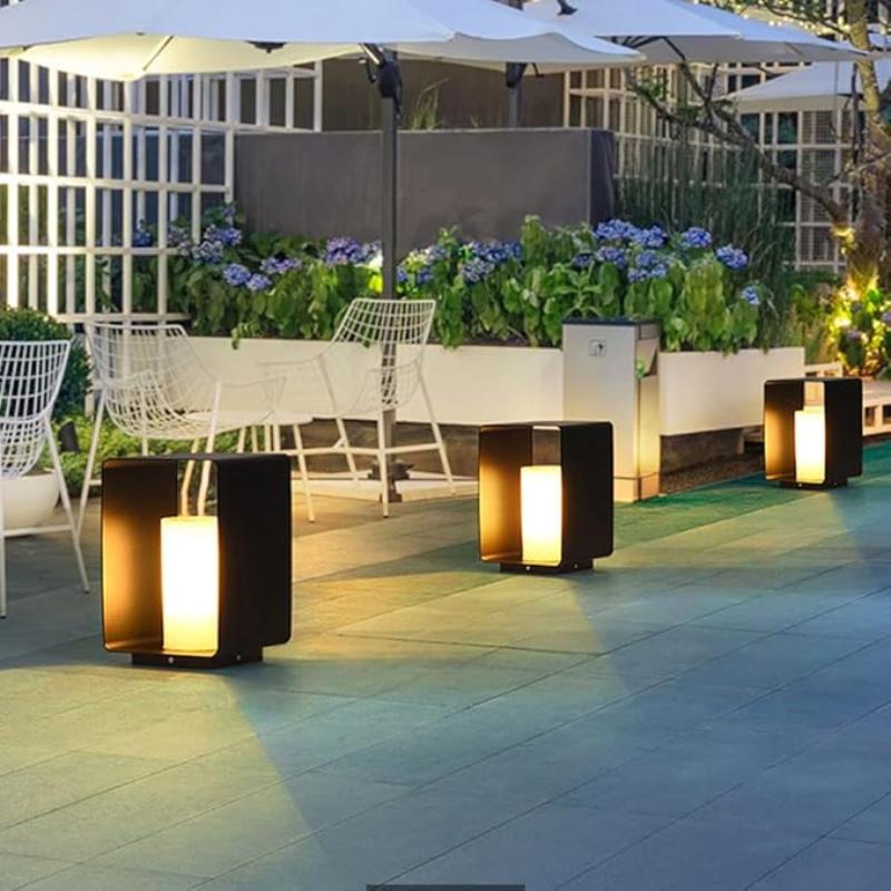 Four modern, dark rectangular outdoor pathway lights with glowing cylindrical diffusers, illuminating a stone patio with white chairs, tables under umbrellas, and planters with blue flowers at dusk.