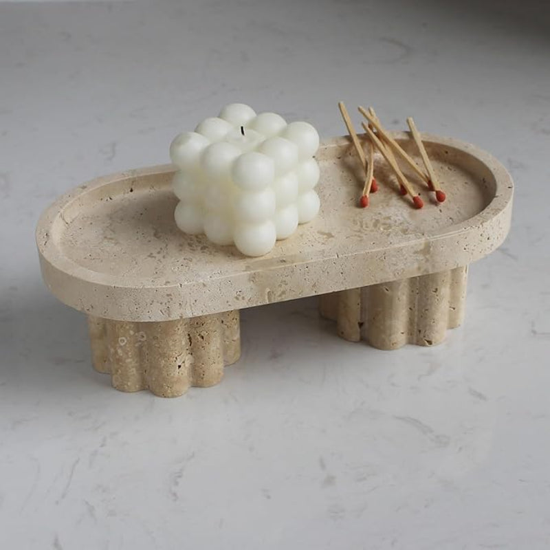 An oval, textured light-colored travertine tray rests on two fluted pedestal legs on a light marble surface. A white bubble candle sits on the left side of the tray, and a small bundle of wooden matches with red tips lies on the right side.