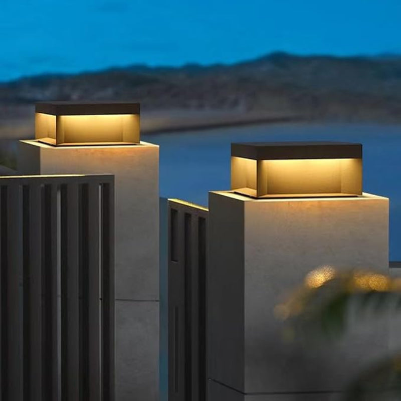 Two modern, rectangular outdoor lights with dark tops and glowing warm-toned panels, mounted on top of gray concrete pillars near a dark fence at twilight.