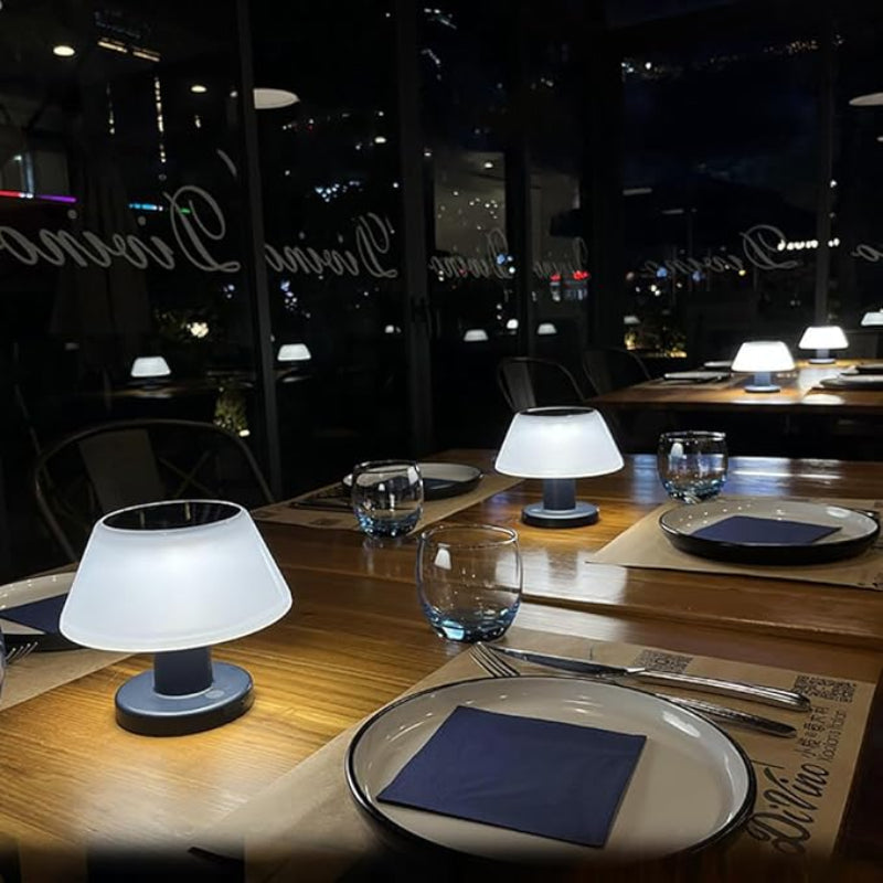 Several small, modern table lamps with white, slightly tapered shades and dark bases illuminate wooden tables set for dining in a dimly lit restaurant at night. Each place setting includes plates with blue napkins, silverware, and drinking glasses. Large windows show the dark exterior with reflections of interior lights.