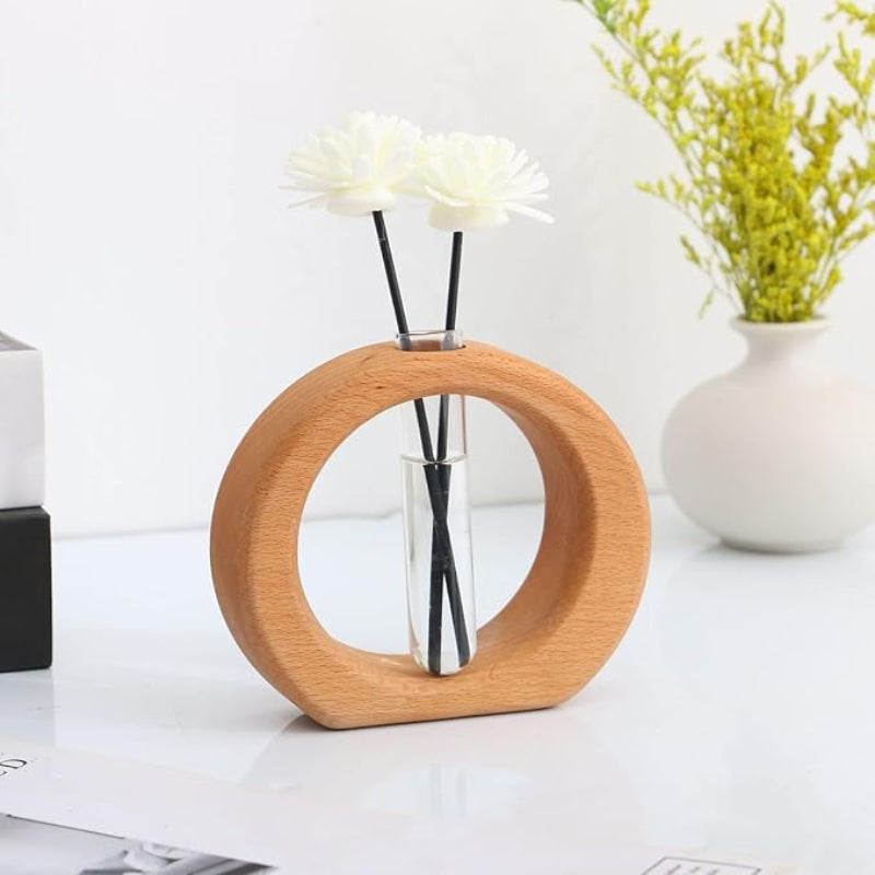 A light wooden circular vase holds a clear glass test tube with two artificial white flowers. A white vase with yellow flowers is visible in the blurred background.