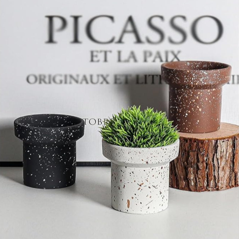 Three small, cylindrical plant pots with slightly flared rims are arranged on a white surface. The pot in the center is white with black and brown speckles and contains a small artificial green plant. To its left is a black pot with white speckles. To its right is a brown pot with white speckles, resting on a small wooden log slice. A book with the word "PICASSO" prominently displayed is in the blurred background.