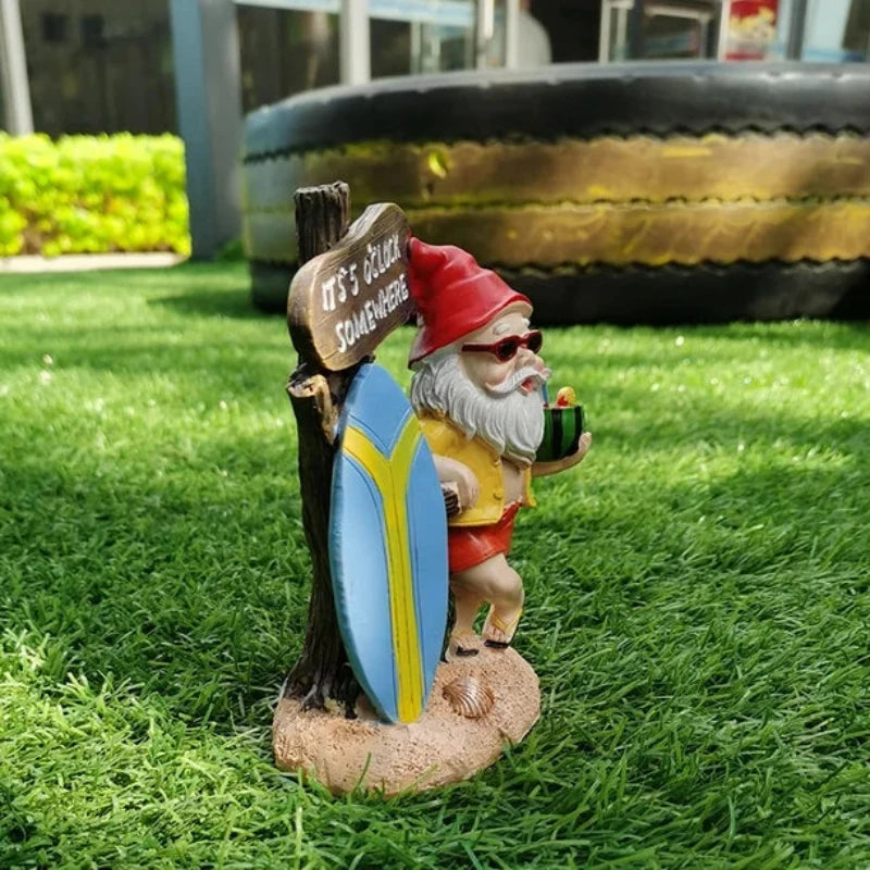 A small, decorative garden gnome figurine with a white beard and sunglasses wears a red pointed hat, a yellow open shirt, and red shorts with black sandals. He holds a green drink in one hand and a small pipe in the other. Behind him is a rustic wooden sign that reads "IT'S 5 O'CLOCK SOMEWHERE," leaning against a blue and yellow surfboard. The gnome stands on a sandy base with small shells, placed on artificial green grass. The blurred background shows outdoor elements like a tire and greenery.