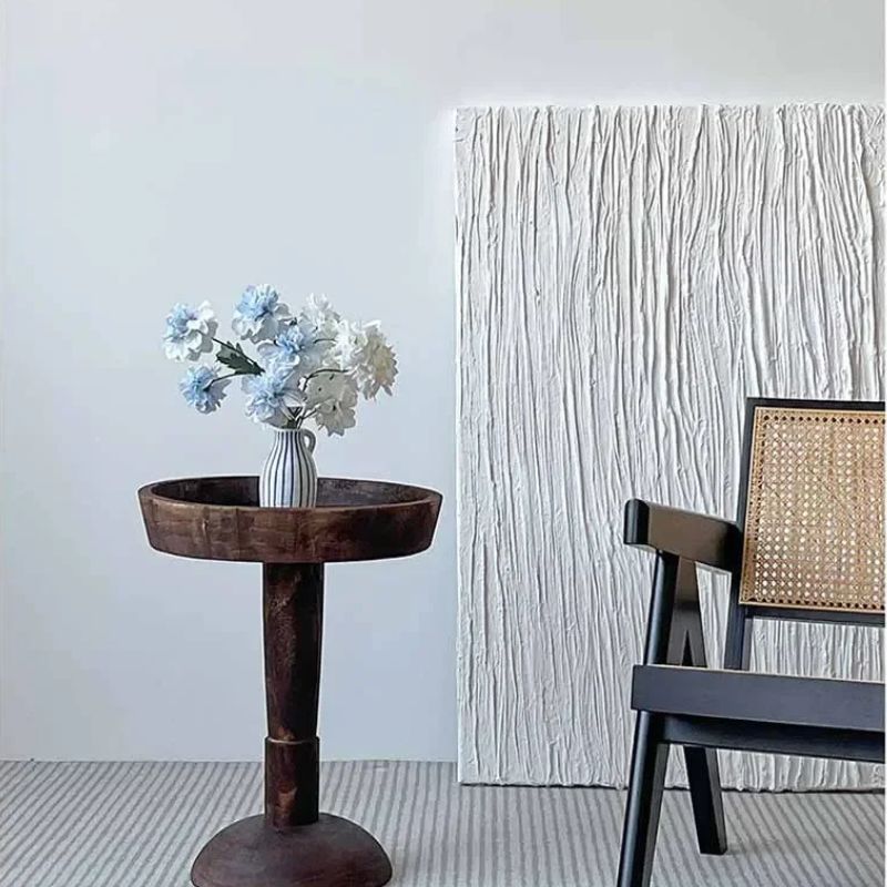 An interior scene with a dark wood, round side table holding a vase of blue and white flowers, a white textured painting on the wall, a black chair with a woven back, and a striped rug.