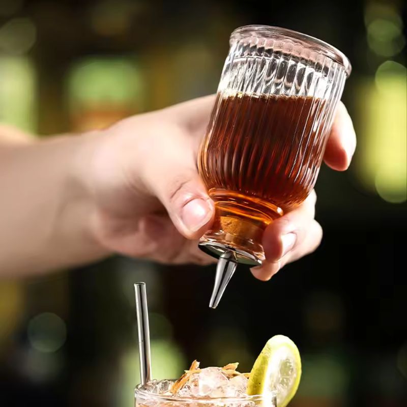 A hand pours a dark amber liquid from a ribbed glass bitters bottle with a metal spout into a cocktail garnished with lime and citrus zest.