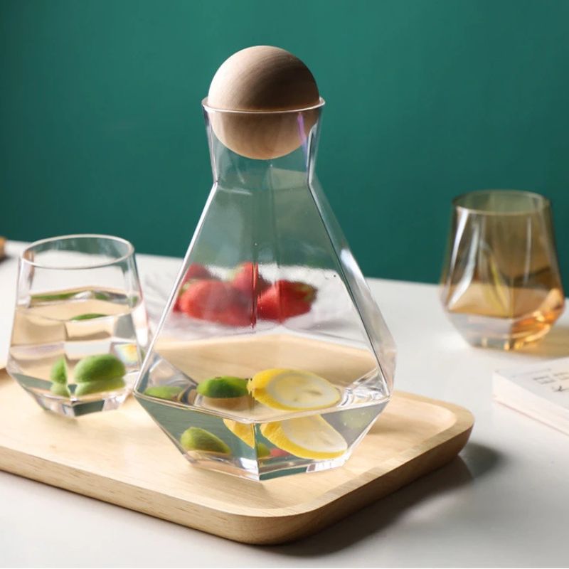Clear glass pitcher and two faceted glasses on a light wooden tray. The pitcher and glasses contain water with fruit slices. The background is a dark teal color.