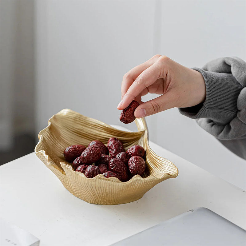 Gold-colored decorative bowl with a textured, sculpted design, filled with dried red dates, placed on a white table with a hand picking one up.