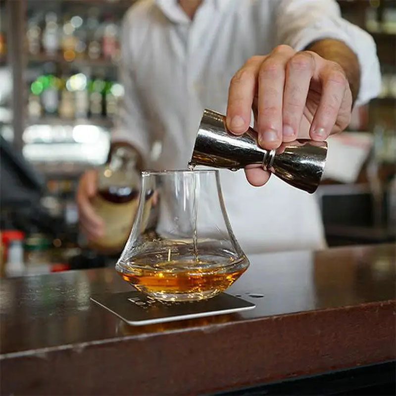 A bartender in a white shirt pouring whiskey into a uniquely shaped glass using a stainless steel jigger, with a well-stocked bar in the background.