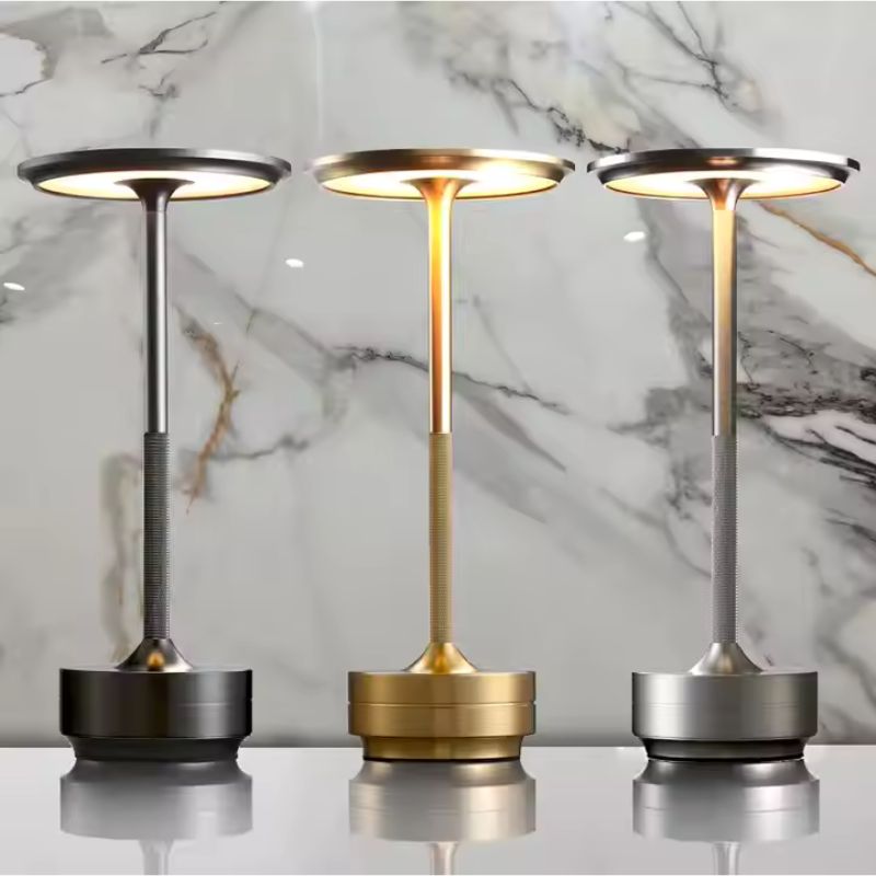 Three modern LED desk lamps in black, gold, and silver, placed side by side on a reflective surface against a marble-patterned background. Each lamp features a circular flat top with warm lighting and a textured cylindrical stem.