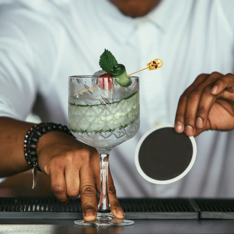 A bartender preparing a cocktail with cucumber, cherry, mint, and a skull-shaped cocktail pick, holding a coaster.
