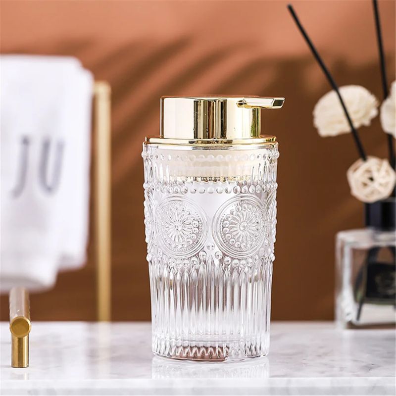Crystal glass soap dispenser with a gold pump on a marble surface, with a white towel showing the letters "JU" in the background.