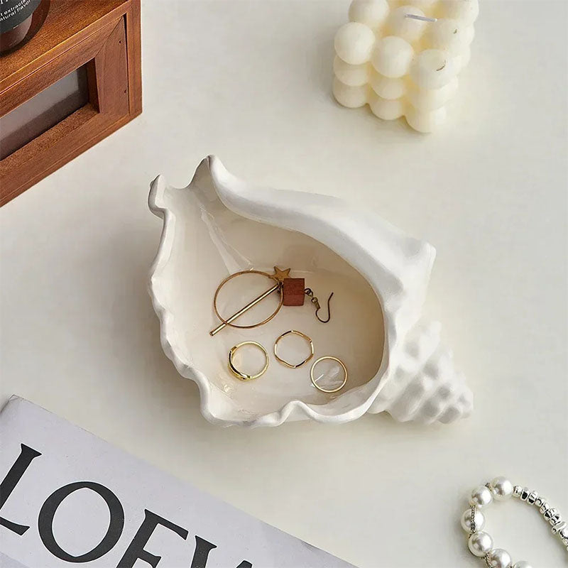 A white ceramic seashell-shaped trinket dish holding gold jewelry, placed on a white surface with a wooden frame, a decorative candle, and a magazine nearby.
