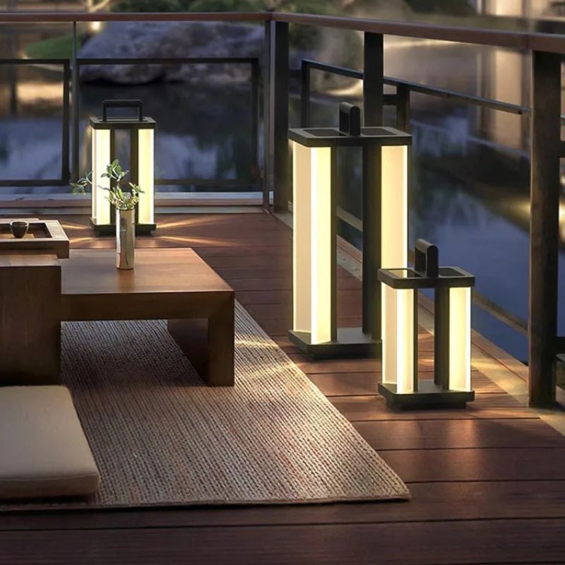 Three illuminated "Villa Solar Lamps" of different sizes stand on a wooden deck near a low table with a vase and a woven rug, overlooking a dark body of water at night.