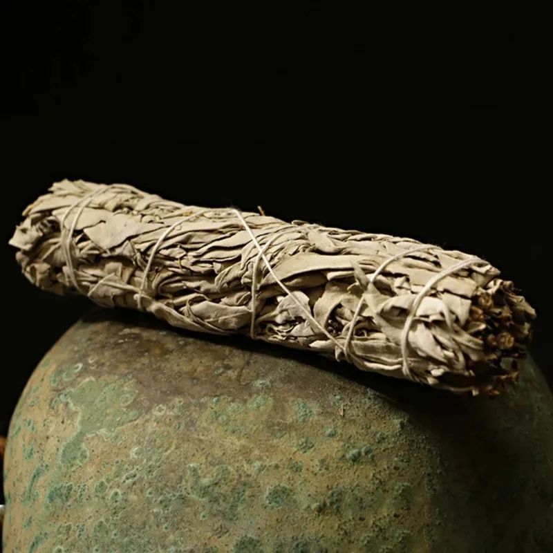 A close-up shot of a thick bundle of dried white sage, tied with white string, resting horizontally on top of a rounded, textured, greenish-bronze object. The sage bundle is the primary focus, showing its pale gray-green leaves and rough texture. The rounded object appears to be a vintage or handcrafted piece, with a mottled green and bronze patina and a rough, uneven surface. The background is a dark, solid color, providing a stark contrast that highlights the sage and the rounded object.