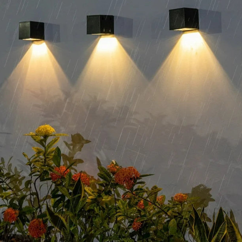 Three black square-shaped solar wall lamps are mounted on a white wall, casting warm yellow light in a downward triangular pattern. The scene is set outdoors in the rain, with raindrops visibly falling. In the foreground, lush green plants with red and yellow flowers are illuminated by the lights.