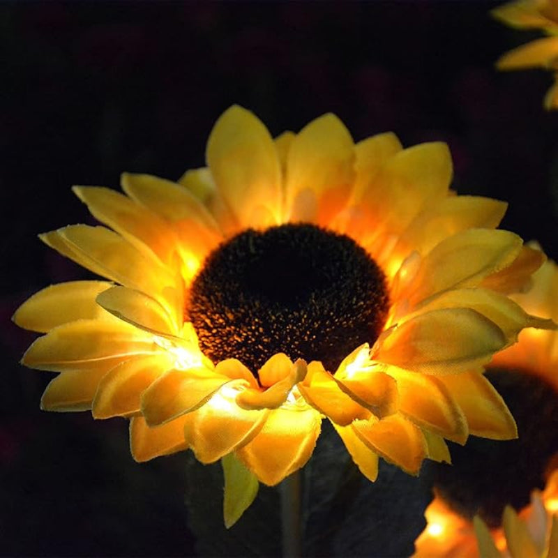 A close-up shows a glowing artificial sunflower at night. The flower has bright yellow petals that are backlit, creating a warm illumination around its dark brown center. Part of another illuminated sunflower is visible on the right against a dark background.