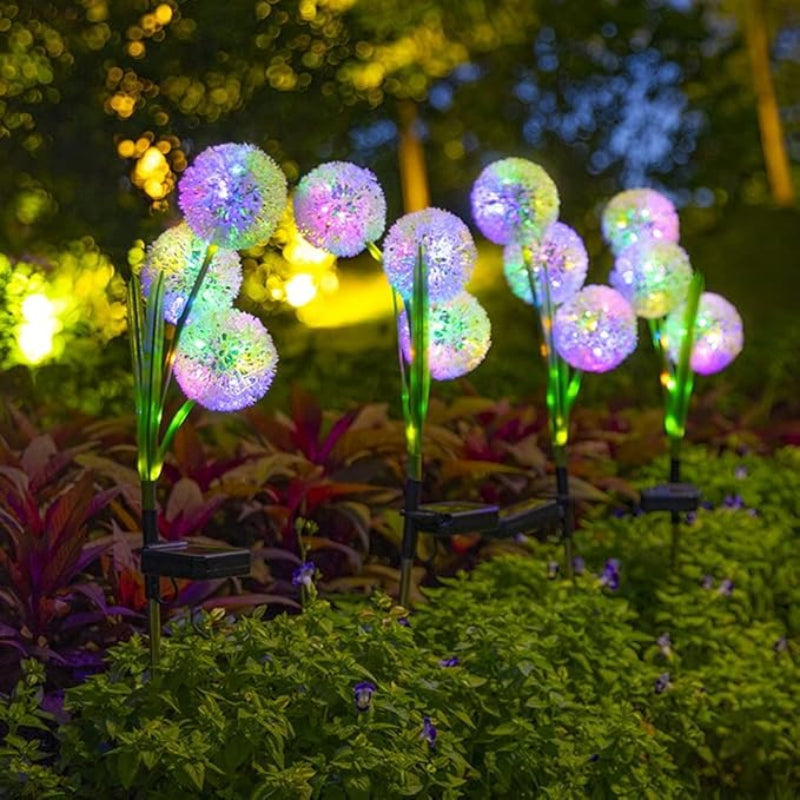The image showcases six solar-powered garden lights, arranged in two rows, resembling dandelions. Each light emits a vibrant, multicolored glow, including pink, yellow, blue, and green. The lights feature green, leaf-shaped accents and black solar panels at their base. They are placed among low-lying greenery and small red flowers, suggesting they are illuminating a garden at night. The background is dark, emphasizing the brightness of the lights.