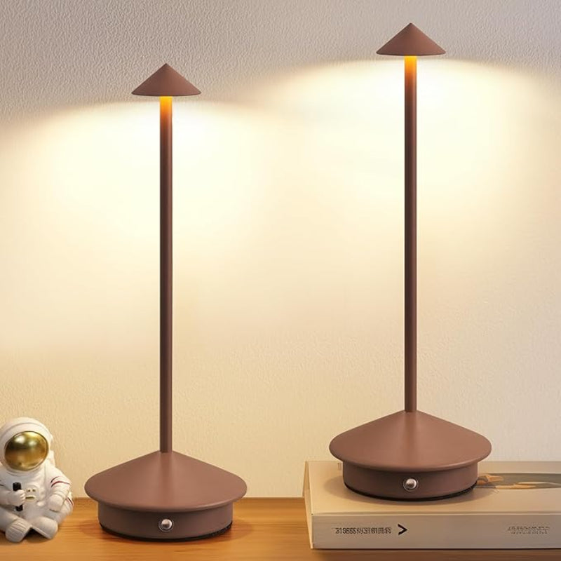 Two illuminated cordless table lamps with slender brown stems and pointed brown shades stand on a light-colored surface. The lamp on the left is next to a small astronaut figurine, and the lamp on the right is placed on a book. A light-colored wall is in the background.