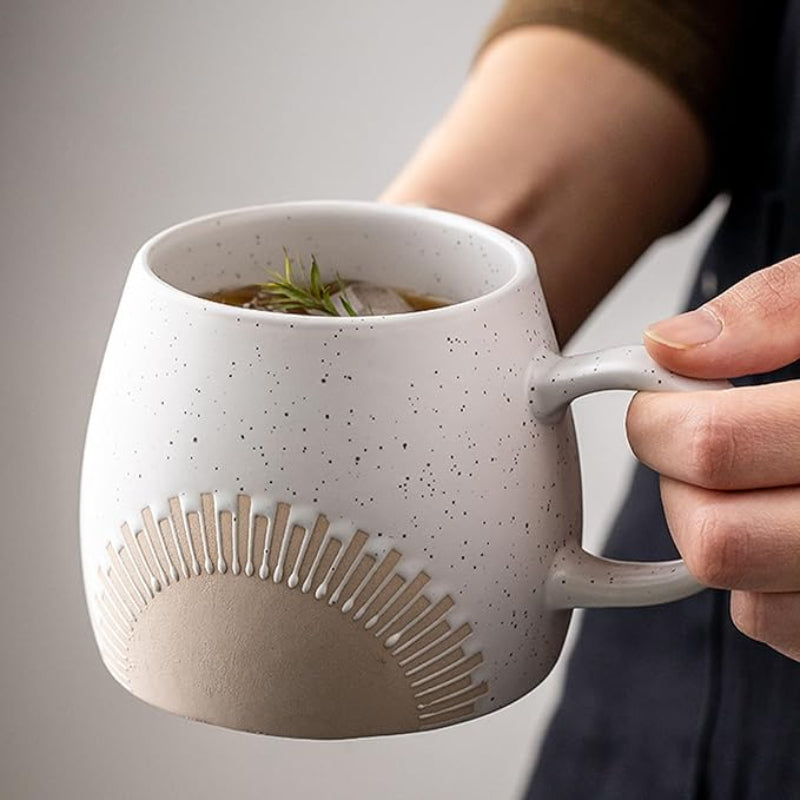 A person holding a speckled ceramic mug with an embossed sunburst design. The mug is filled with a beverage, ice cubes, and a sprig of greenery, giving it a refreshing appearance.