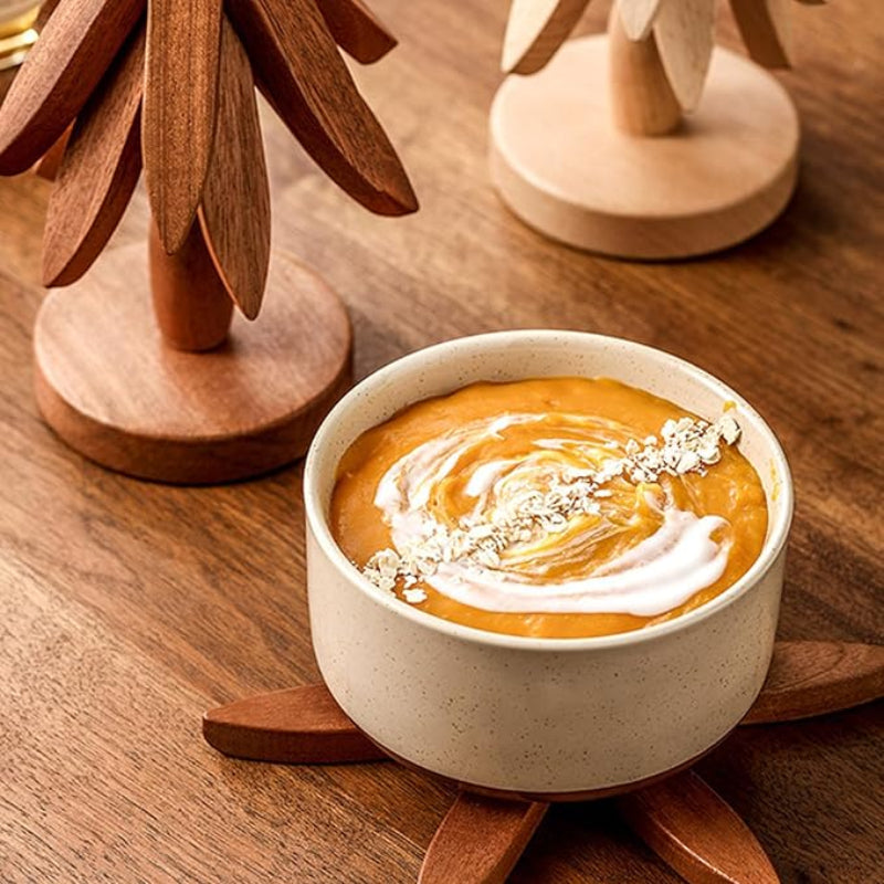 A close-up shot shows a light-colored bowl of orange soup with a swirl of white cream and a sprinkle of white flakes on a starburst-shaped wooden trivet. Two tree-shaped wooden trivets stand in the background.