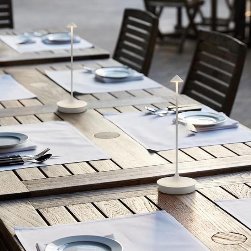 Several outdoor dining tables are set with white placemats, plates, silverware, and illuminated cordless lamps with slender stems and pointed shades. Wooden chairs are placed around the tables. The setting suggests a restaurant patio at dusk or dawn.