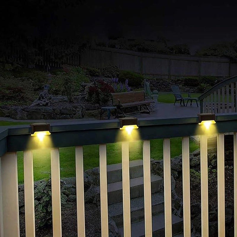 Three small, dark, rectangular lights are mounted on a white wooden railing at night, casting a warm yellow downward glow. The railing overlooks a stone staircase leading to a garden with greenery, a bench, and chairs. A dark fence is visible in the background.