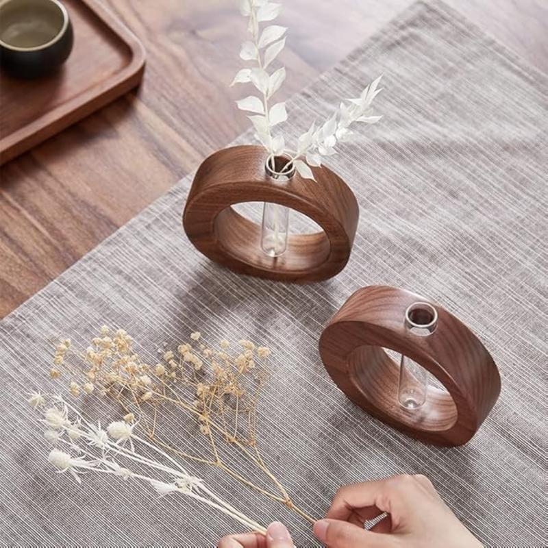 Two dark wooden circular vases, each holding a clear glass test tube, sit on a textured gray surface. One vase contains dried white flowers, while a hand holds a bundle of dried, delicate flowers near the other vase. A small cup on a wooden tray is visible in the upper left corner.