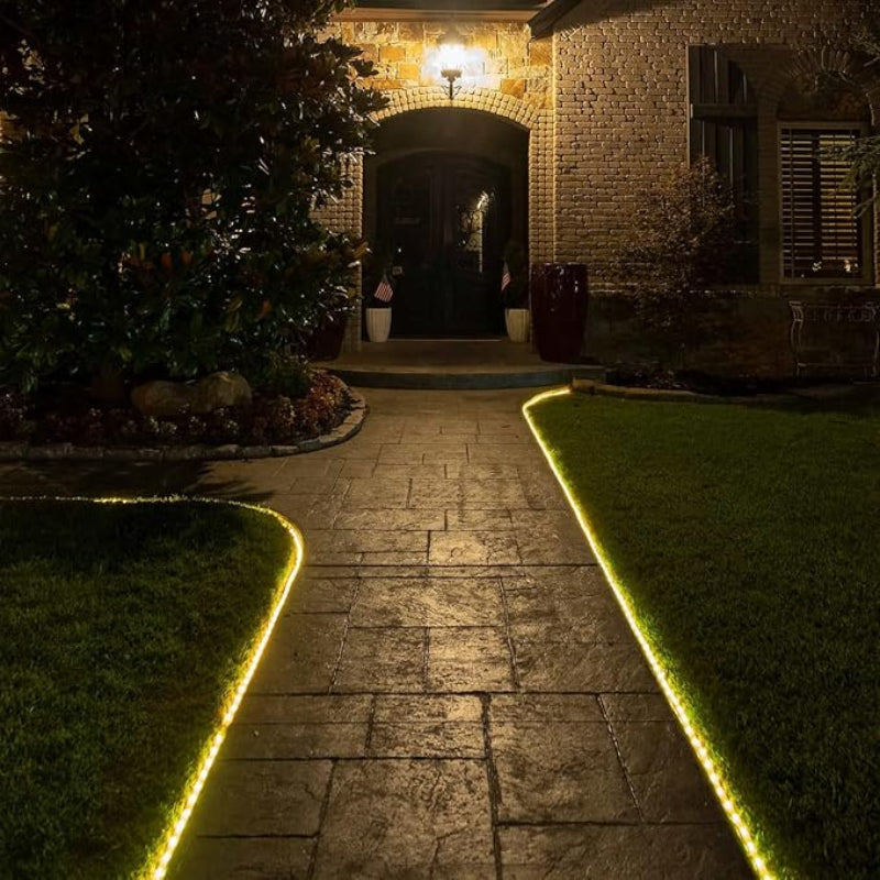 A warm white rope light outlines a curved stone walkway leading to the front door of a brick house at night. The rope light illuminates the edges of the path, which is bordered by green grass. A porch light above the arched doorway provides additional illumination.