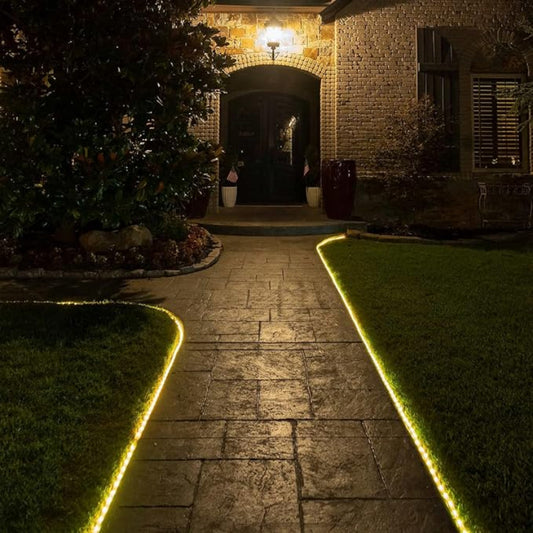 A warm white rope light outlines a curved stone walkway leading to the front door of a brick house at night. The rope light illuminates the edges of the path, which is bordered by green grass. A porch light above the arched doorway provides additional illumination.